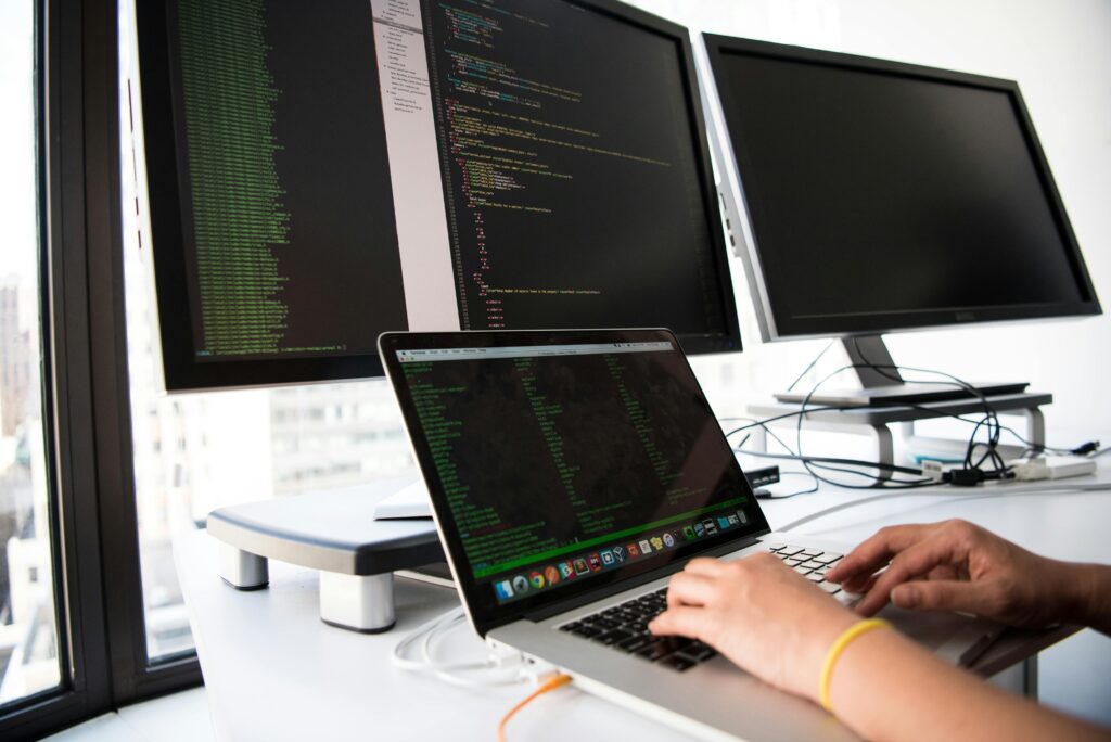 A developer writes code on a laptop in front of multiple monitors in an office setting.